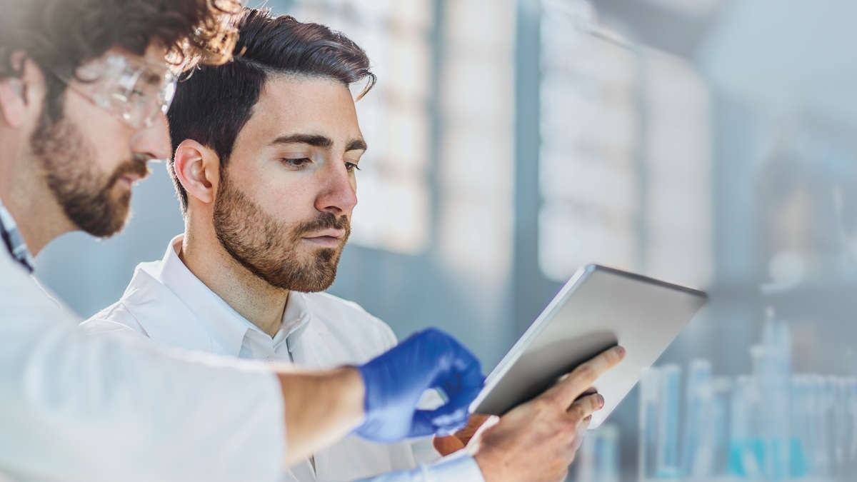 Two scientist looking on a tablet in their lab, discussing, pointing, one wearing glasses and gloves
